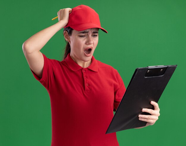 Jeune Femme De Livraison En Uniforme Et Casquette Tenant Un Presse-papiers Et Un Crayon Regardant Le Presse-papiers Mettant La Main Sur La Tête Isolée Sur Le Mur