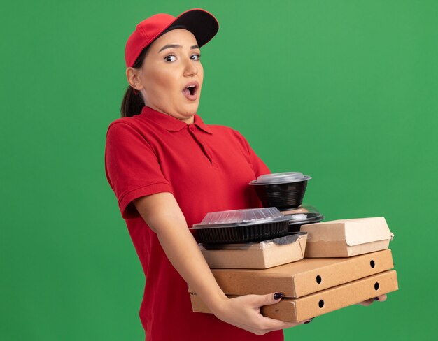 Jeune femme de livraison en uniforme rouge et cap tenant des boîtes de pizza et des emballages alimentaires à l'avant étonné et surpris debout sur le mur vert