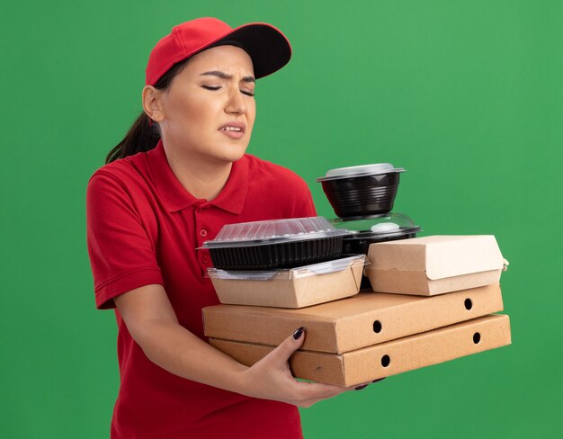 Jeune femme de livraison en uniforme rouge et cap tenant des boîtes de pizza et des emballages alimentaires à la confusion et mécontent debout sur le mur vert