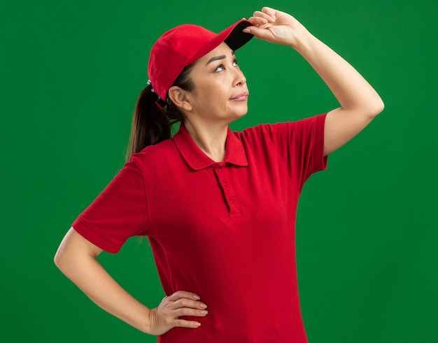 Jeune femme de livraison en uniforme rouge et casquette en levant avec une expression sceptique