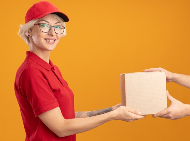 Jeune femme de livraison en uniforme rouge et casquette portant des lunettes donnant une boîte en carton à u client souriant sympathique debout sur le mur orange