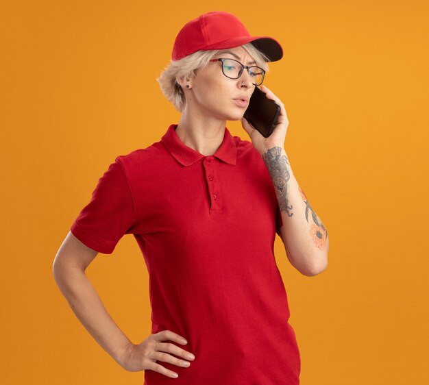Jeune femme de livraison en uniforme rouge et casquette portant des lunettes parler au téléphone mobile avec un visage sérieux debout sur un mur orange