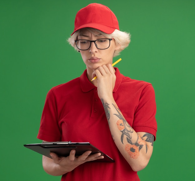 Jeune femme de livraison en uniforme rouge et casquette portant des lunettes avec presse-papiers avec des pages blanches et un crayon regardant vers le bas avec un visage sérieux pensant debout sur le mur vert