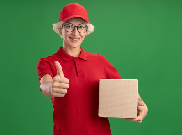 Jeune femme de livraison en uniforme rouge et casquette portant des lunettes tenant une boîte en carton à montrant les pouces vers le haut souriant joyeusement debout sur le mur vert