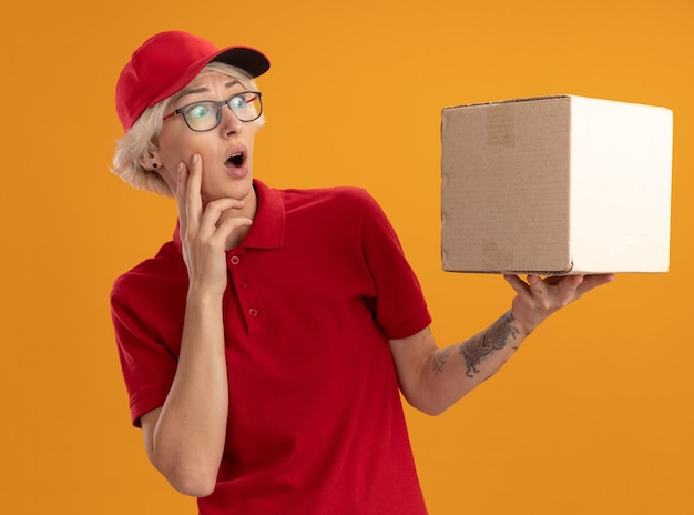 Jeune femme de livraison en uniforme rouge et casquette portant des lunettes tenant une boîte en carton à la regarder étonné et surpris debout sur un mur orange