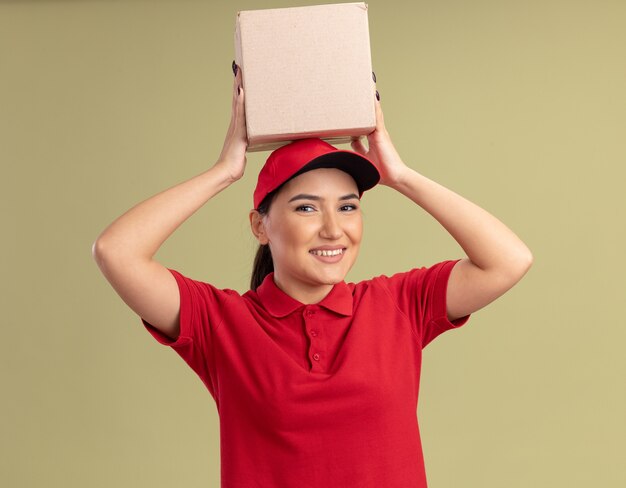 Jeune femme de livraison en uniforme rouge et casquette tenant une boîte en carton sur sa tête à l'avant souriant joyeusement debout sur le mur vert