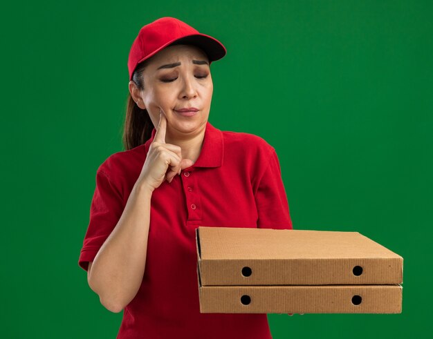 Jeune femme de livraison en uniforme rouge et casquette tenant des boîtes à pizza les regardant être confondues