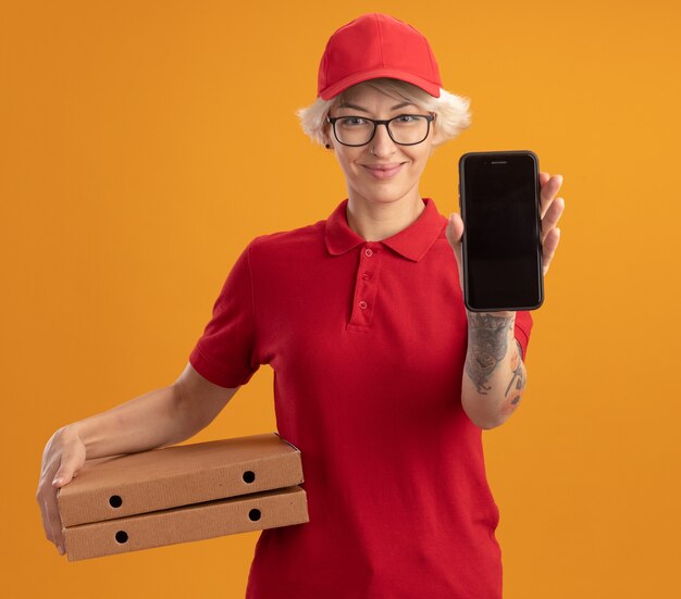 Jeune femme de livraison en uniforme rouge et chapeau portant des lunettes montrant smartphone tenant des boîtes à pizza souriant joyeusement debout sur un mur orange