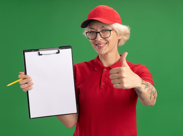 Photo gratuite jeune femme de livraison en uniforme rouge et chapeau portant des lunettes avec presse-papiers avec des pages blanches et un crayon souriant joyeusement montrant les pouces vers le haut debout sur le mur vert