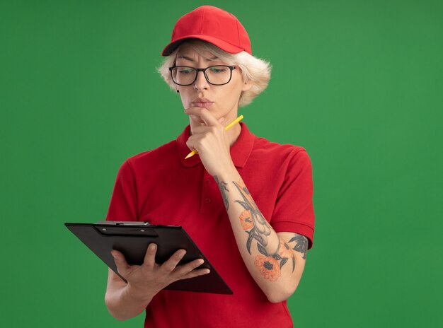 Jeune femme de livraison en uniforme rouge et chapeau portant des lunettes tenant le presse-papiers et un crayon à la recherche avec un visage sérieux pensant debout sur le mur vert