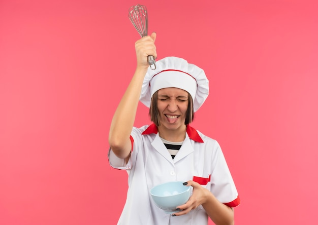 Photo gratuite jeune femme ludique cuisinier en uniforme de chef tenant le bol et élever le fouet au-dessus de la tête et montrant la langue avec les yeux fermés isolé sur fond rose avec copie