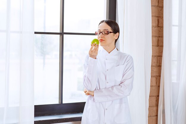 Jeune femme avec des lunettes en blouse de laboratoire mangeant une pomme verte près de la fenêtre.