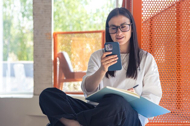 Une jeune femme à lunettes avec un smartphone avec des blocs-notes dans un intérieur moderne