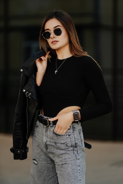 Jeune femme à lunettes de soleil, chapeau et veste en cuir noir posant en plein air.