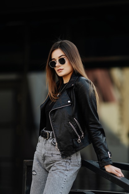Jeune femme à lunettes de soleil et veste en cuir noir posant en plein air