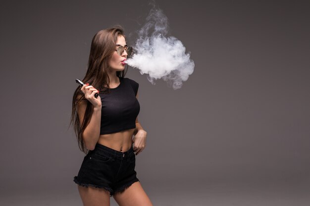 Jeune femme à lunettes de soleil et vêtements noirs vaping et souffle de la fumée de cigarette électronique sur mur gris