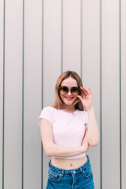 Jeune femme en lunettes de soleil vintage dans des vêtements de marque élégant près d'un mur à rayures métalliques