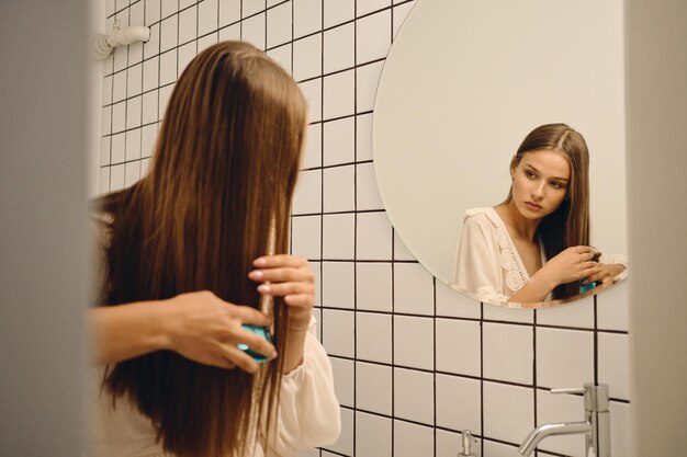 Jeune femme magnifique en robe blanche se brossant pensivement les cheveux près du miroir dans la salle de bain seule