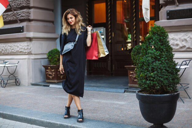 Jeune femme magnifique en robe noire avec sac croisé regardant rêveusement de côté tenant des sacs à provisions colorés à la main tout en marchant sur la belle rue confortable de la ville seule