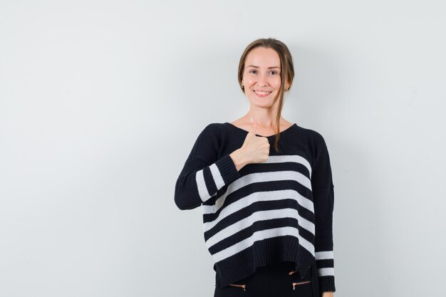 Jeune femme en maille rayée et pantalon noir montrant le pouce vers le haut et à la heureux