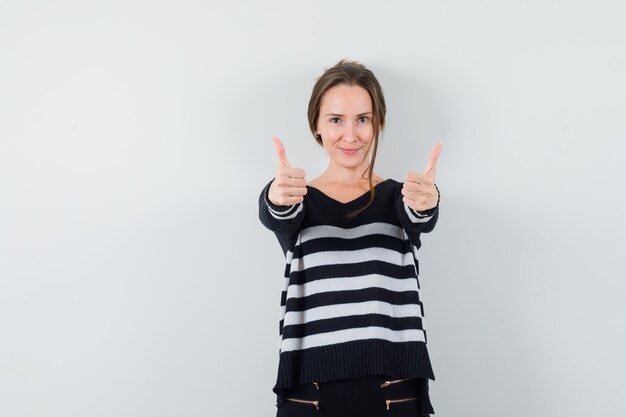 Jeune femme en maille rayée et pantalon noir montrant les pouces vers le haut et à heureux