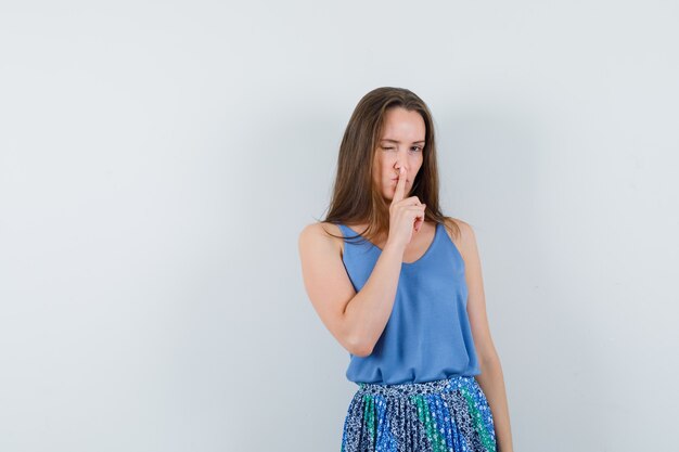 Jeune femme en maillot, jupe montrant le geste de silence et l'air confiant, vue de face.