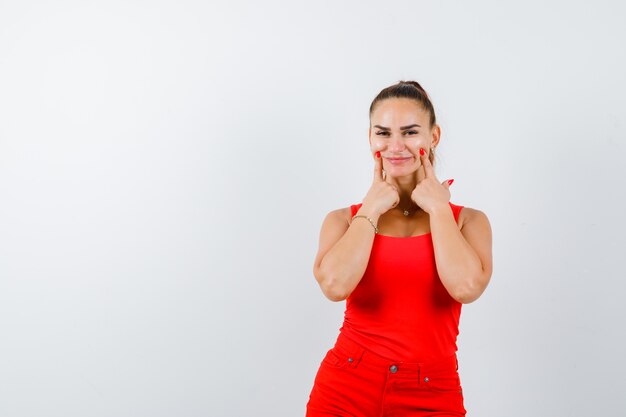 Jeune femme en maillot rouge, pantalon rouge appuyant sur les joues avec les doigts et à la joyeuse vue de face.