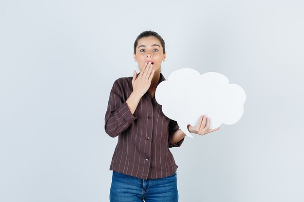 Jeune femme avec la main sur la bouche, gardant l'affiche en papier en chemise, jeans et l'air surpris, vue de face.