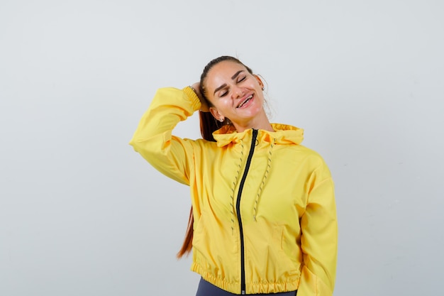 Jeune femme avec la main derrière la tête en veste jaune et charmante, vue de face.