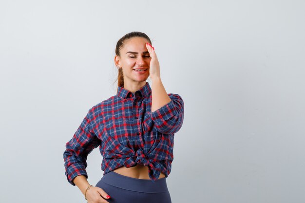 Jeune femme avec la main sur le front en chemise à carreaux, pantalon et l'air heureux, vue de face.