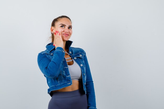 Jeune femme avec la main sur la joue en crop top, veste, pantalon et jolie. vue de face.