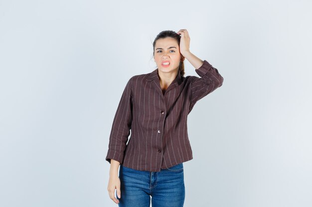 Jeune femme avec la main sur la tête, grimaçant en chemise rayée, jeans et semblant agitée. vue de face.