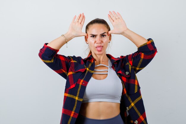 Jeune femme avec les mains au-dessus de la tête comme oreilles en haut court, chemise à carreaux, pantalon et semblant drôle, vue de face.