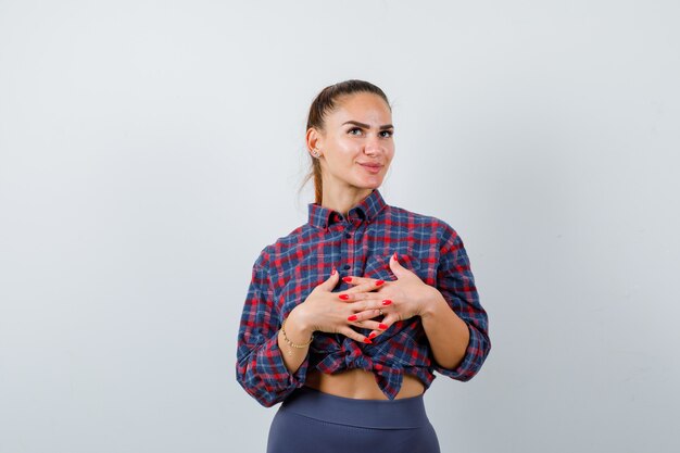 Jeune femme avec les mains avec les doigts entrelacés devant elle en chemise à carreaux, pantalon et à la pensive. vue de face.