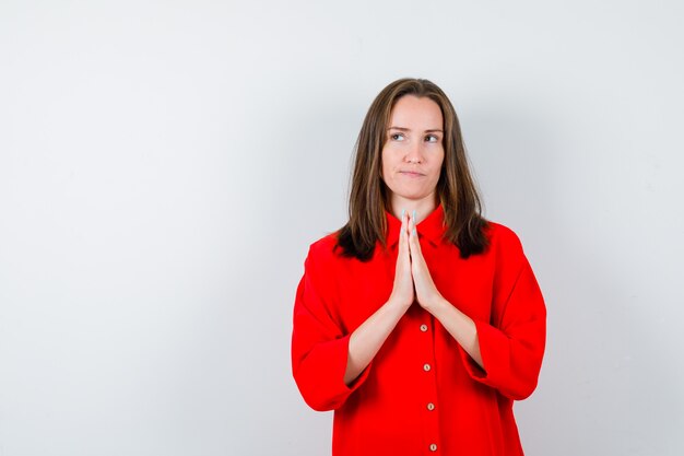 Jeune femme avec les mains en geste de prière en chemisier rouge et l'air pensif. vue de face.