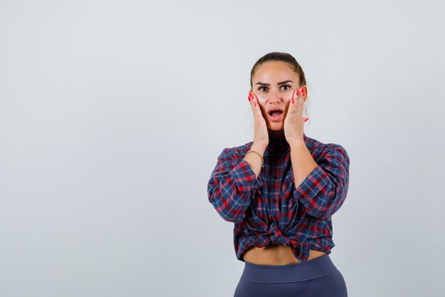 Jeune femme avec les mains sur les joues en chemise à carreaux, pantalon et à la perplexité, vue de face.