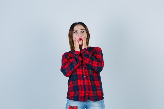 Jeune femme avec les mains près de la bouche, gardant la bouche ouverte dans une chemise à carreaux, un jean et l'air surpris. vue de face.