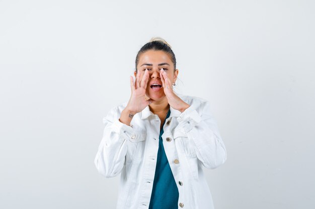 Jeune femme avec les mains près de la bouche pour dire le secret en chemise, veste blanche et semblant excitée, vue de face.