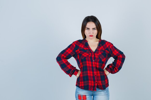 Jeune femme avec les mains sur la taille en chemise à carreaux, jeans et regardant en colère, vue de face.