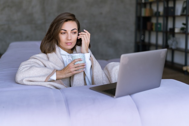 Jeune femme à la maison sur le canapé en hiver sous une couverture confortable avec un ordinateur portable, regardant un film