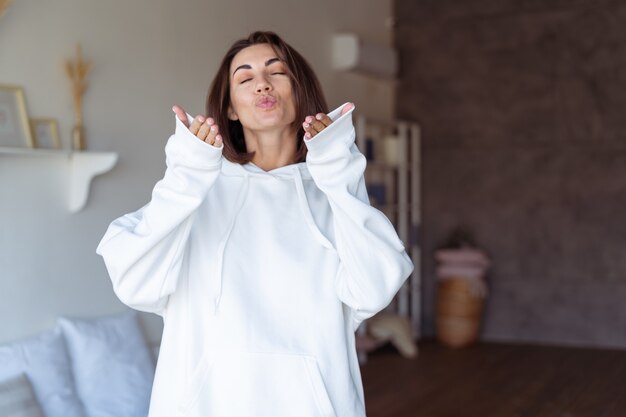 Jeune femme à la maison dans la chambre dans un sweat à capuche blanc chaud, heureuse, posant, souriante, soirée d'hiver confortable