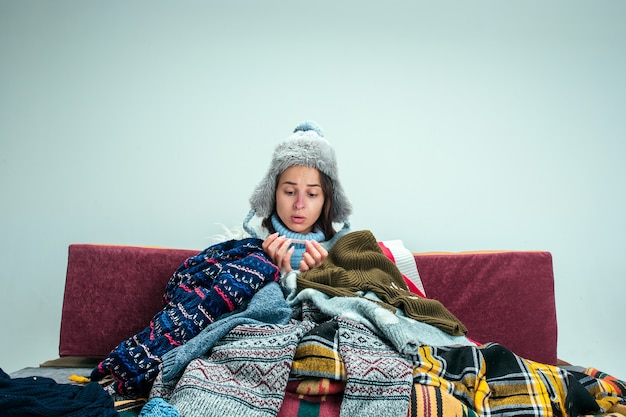 La jeune femme malade avec cheminée assise sur un canapé à la maison ou en studio recouvert de vêtements chauds tricotés. Maladie, grippe, concept de douleur. Détente à la maison. Concepts de santé.