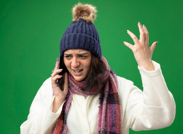 Jeune femme malade en colère portant chapeau d'hiver robe et écharpe parler au téléphone en gardant la main dans l'air à côté isolé sur mur vert