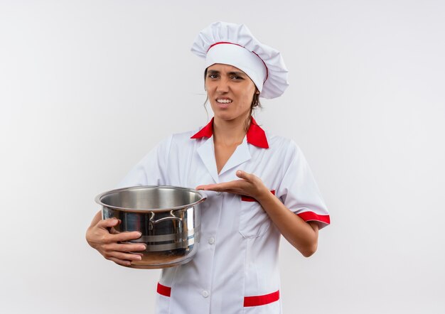 Jeune femme malheureuse cuisinier portant un uniforme de chef tenant et points avec la main à la casserole sur un mur blanc isolé avec copie espace