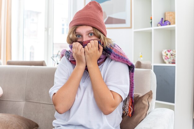 Jeune femme malsaine au chapeau avec une écharpe chaude autour du cou se sentant mal et malade souffrant de rhume et de grippe l'air inquiet assis sur un canapé dans un salon lumineux