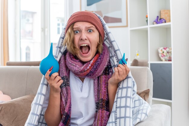 Jeune femme malsaine au chapeau enveloppé dans une couverture, se sentant mal et malade tenant des lavements criant avec une expression agressive agacée et irritée assise sur un canapé dans un salon lumineux