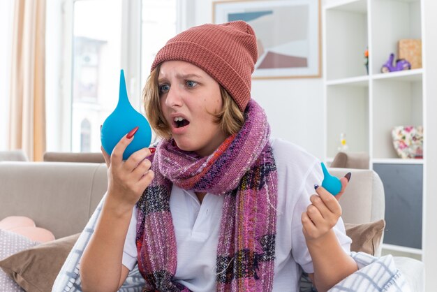 Jeune femme malsaine au chapeau enveloppé dans une couverture se sentant mal et malade tenant des lavements semblant confus ayant des doutes assis sur un canapé dans un salon lumineux