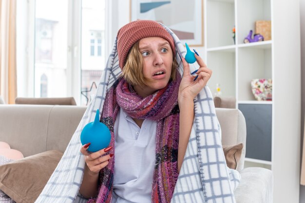 Jeune femme malsaine au chapeau enveloppé dans une couverture se sentant mal et malade tenant des lavements semblant confus ayant des doutes assis sur un canapé dans un salon lumineux