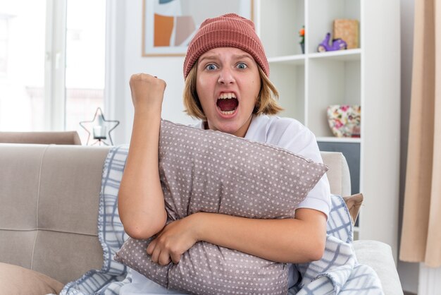 Jeune femme malsaine en colère dans un chapeau chaud avec une couverture semblant malade souffrant du rhume et de la grippe tenant un oreiller serrant le poing criant avec une expression agressive assise sur un canapé dans un salon lumineux