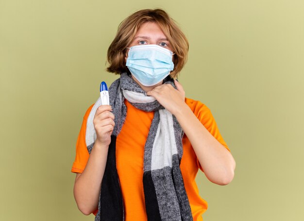 Jeune femme malsaine avec une écharpe chaude autour du cou portant un masque facial protecteur tenant un thermomètre souffrant de maux de gorge touchant le cou debout sur un mur vert